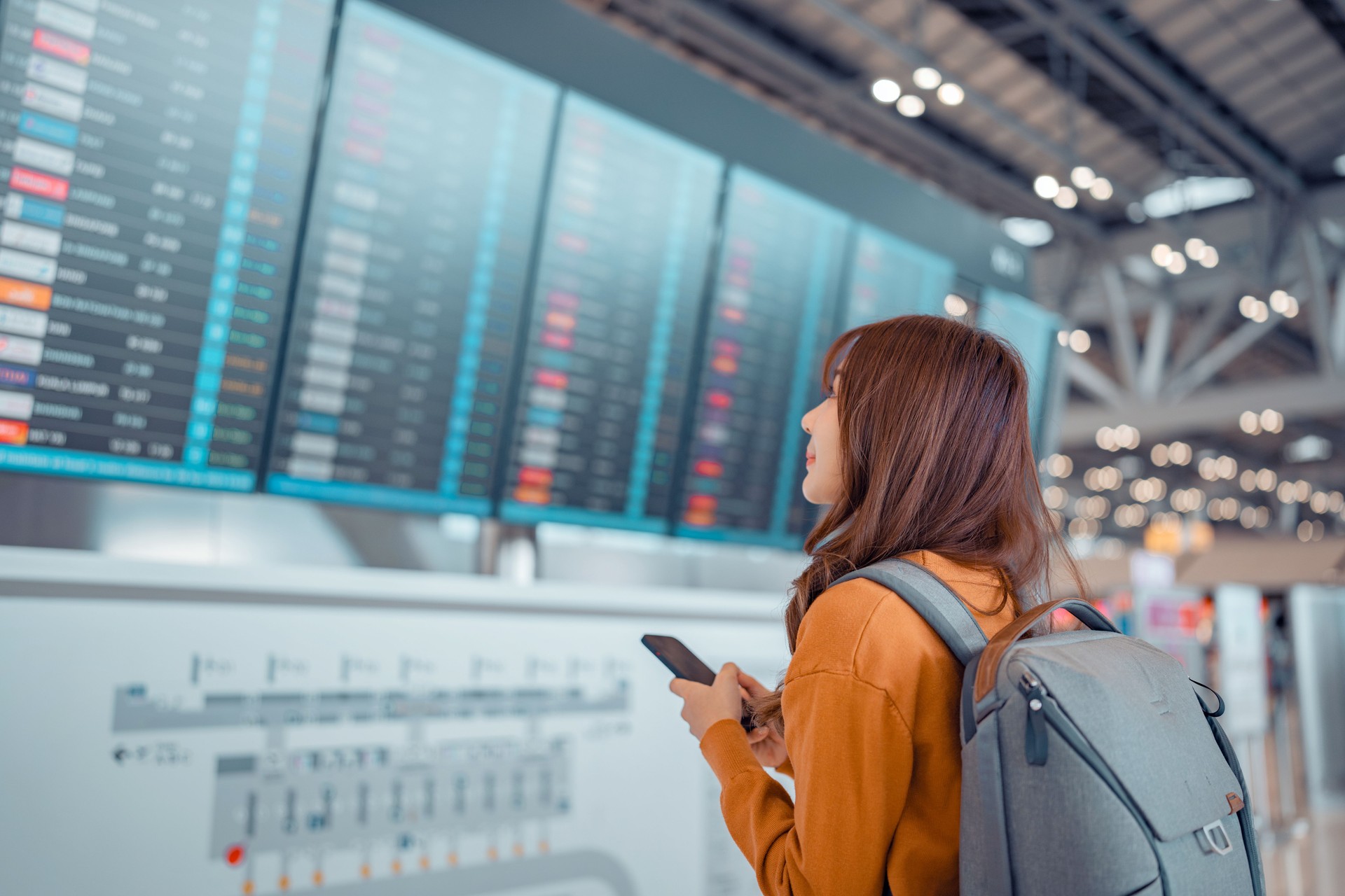 Happy asian woman traveller checking flight schedule departures board in airport terminal hall in front of check in counters. Tourist journey trip concept