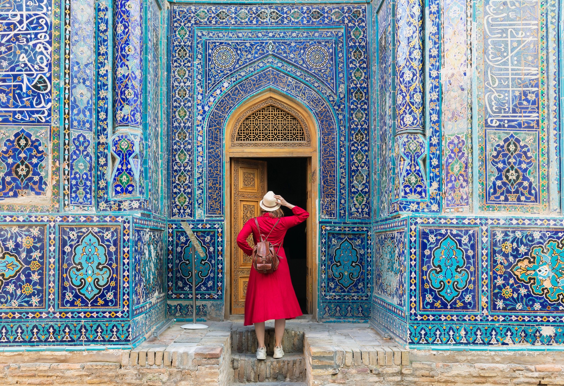 tourist woman in red dress clothes stands near an ancient architectural monument with wooden doors in oriental style with Islamic ornaments in Samarkand Uzbekistan Central Asia